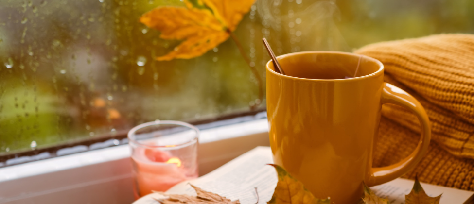 Steaming tea beside book and candle on rainy windowsill